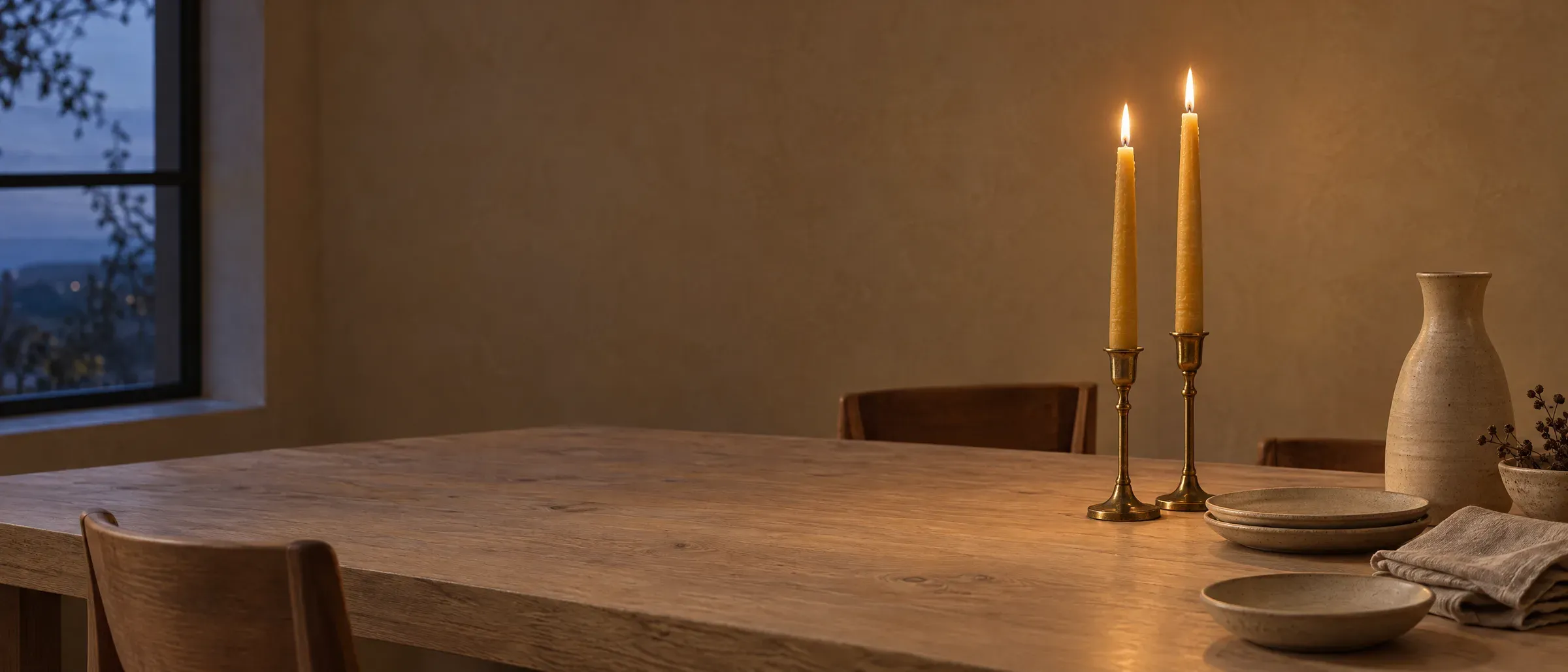 Two beeswax tapers lit in brass candlesticks on an oak dining table